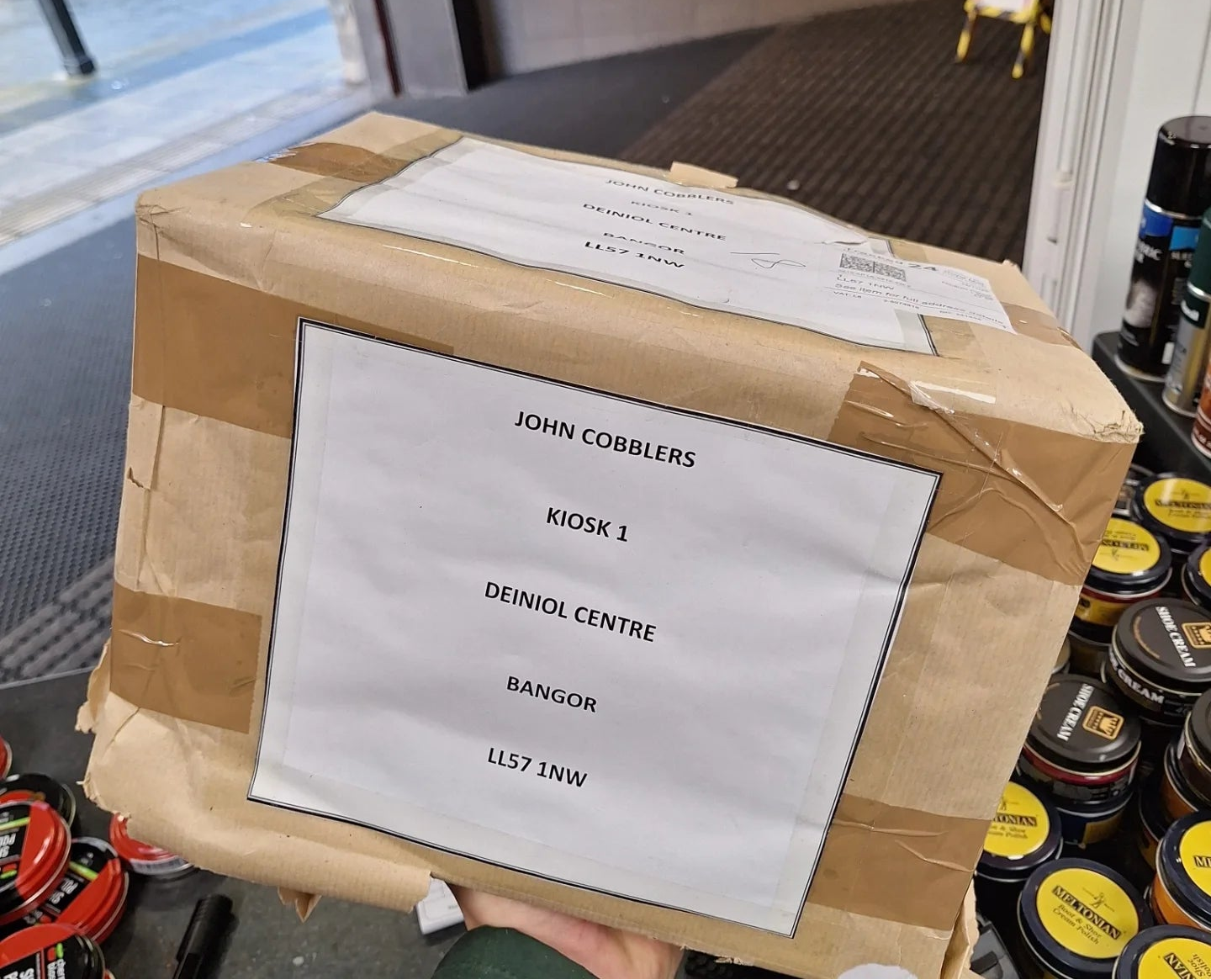 A large brown cardboard box with tape and a white sign reading 'JOHN COBBLERS KIOSK 1 DEINIOL CENTRE BANGOR LLS7 1NW' being held in front of a store entrance. Shiny black shoe polish tins are visible on the right side.